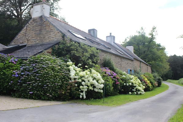 A stone building with a sloped roof, surrounded by lush greenery and flowering plants.