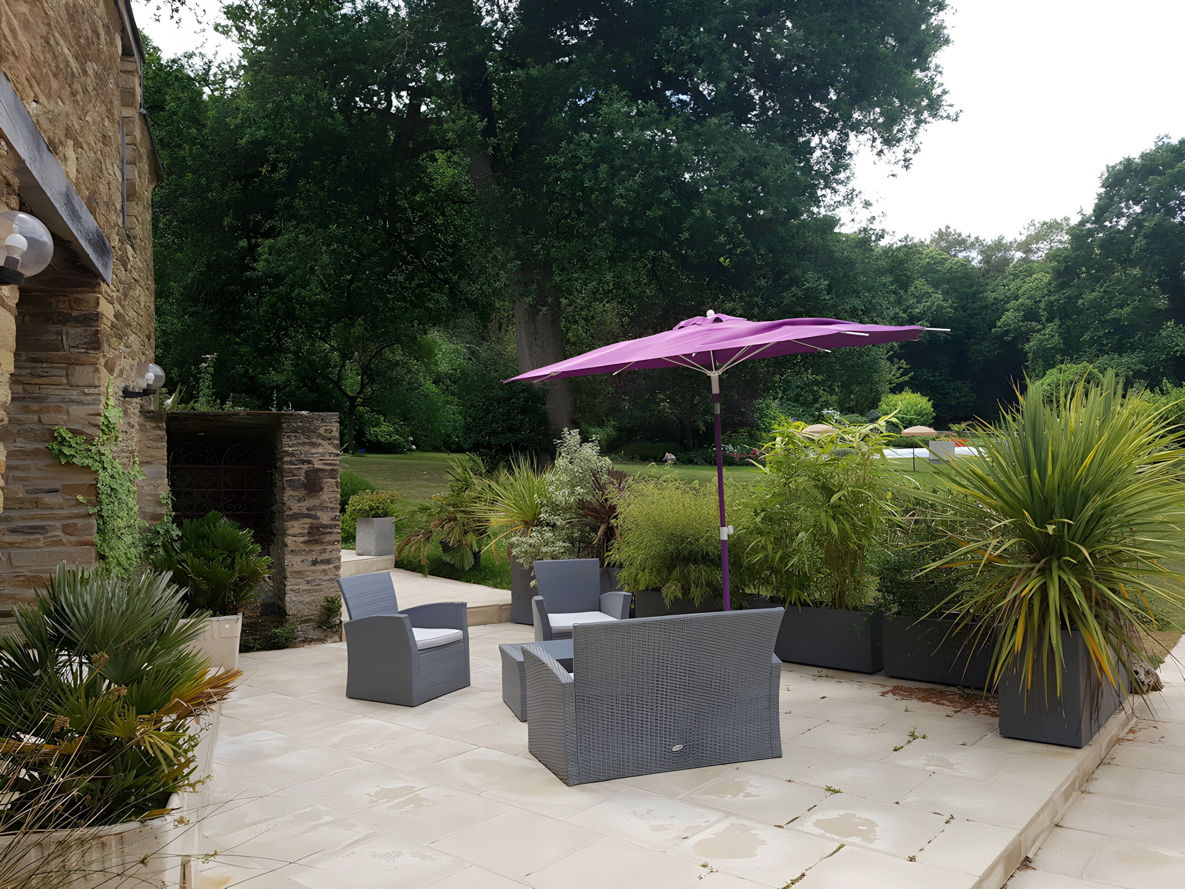 Une terrasse extérieure aménagée avec des meubles en osier et un parasol violet, entourée de verdure et d’un mur en pierre.