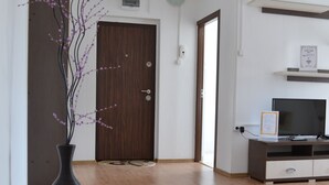 Ein Flur mit Holzboden, einer dunklen Holztür, einer weißen Wand, einem TV-Schrank mit einem Flachbildfernseher und einer Vase mit Zweigen.