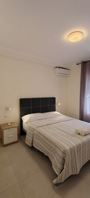 Ett hotellrum med en säng, ett sängbord, en nattdukslampa, en luftkonditioneringsanläggning och ett fönster med gardiner.