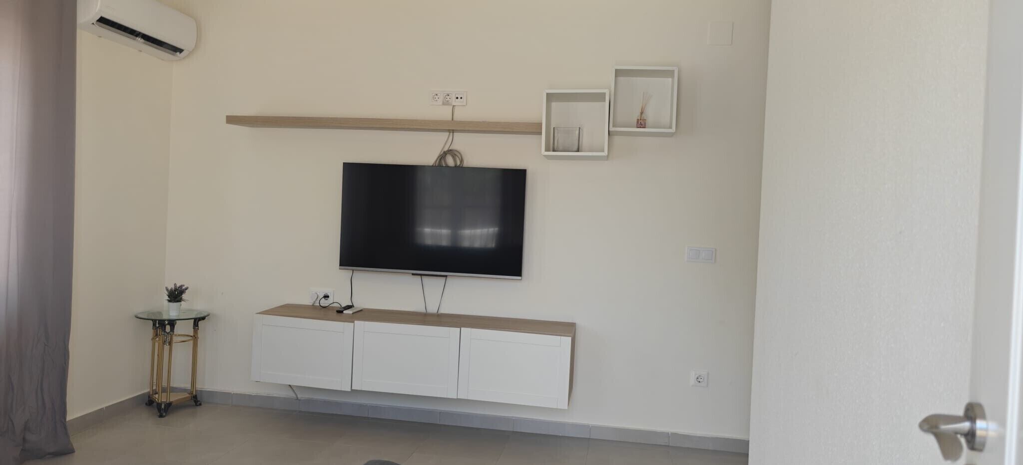 Quarto com televisão na parede, uma mesinha lateral com uma planta, um armário branco e uma estante com objetos decorativos.