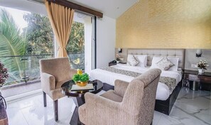 Una habitación de hotel moderna con una cama grande, dos sillones, una mesita con una planta y un balcón con vistas a zonas verdes.