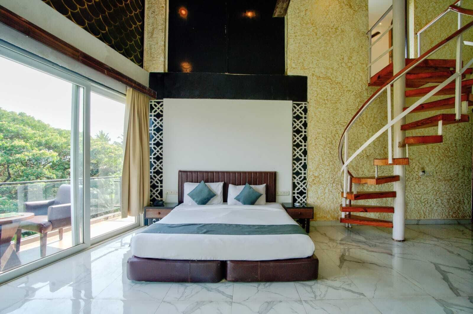 A bedroom with a large bed, a nightstand, a chair, a glass table, a staircase, and a view of trees.