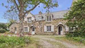 Une maison en pierre avec une allée gravillonnée, un grand arbre et un jardin fleuri.