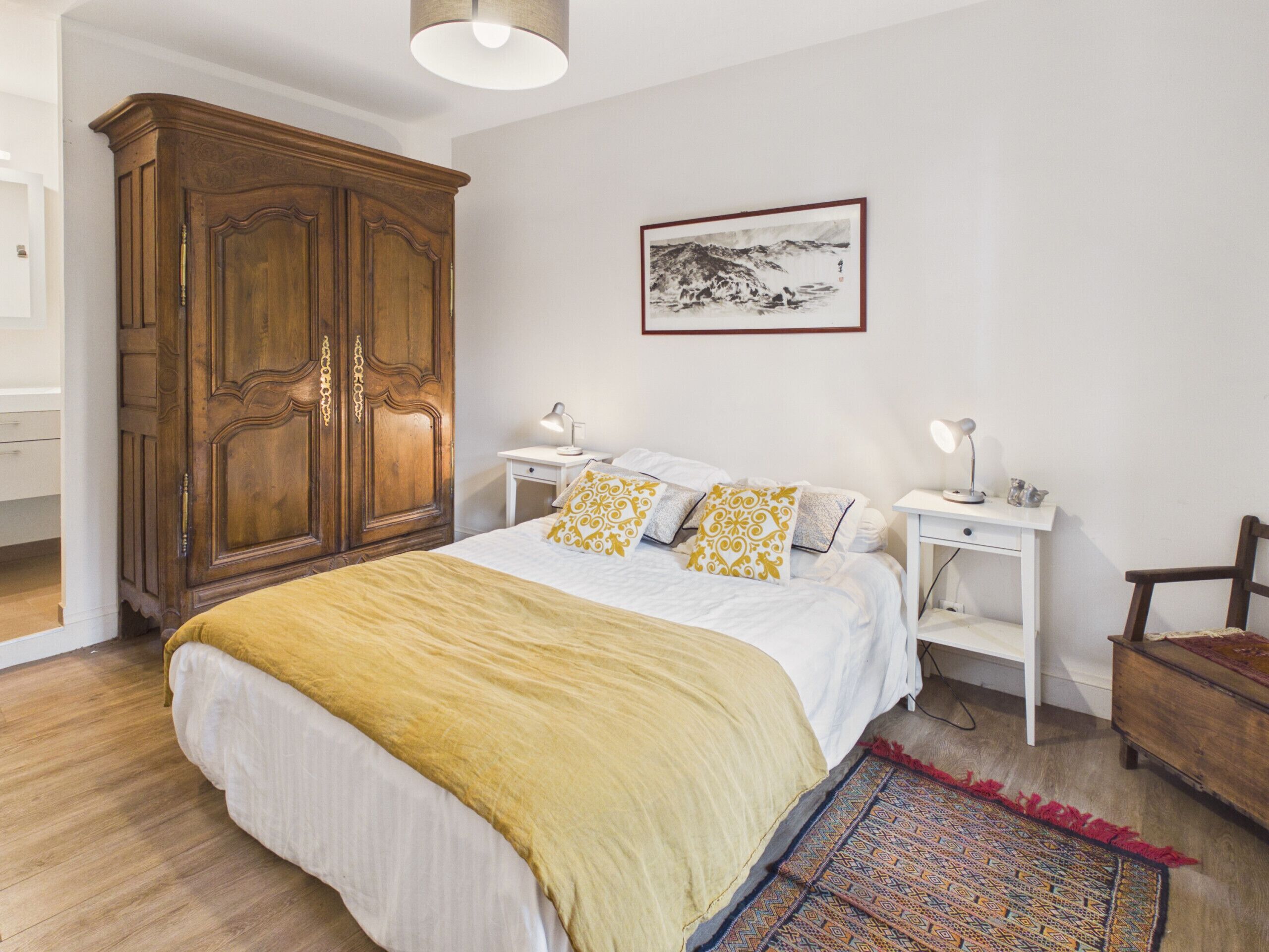 A bedroom with a large wooden wardrobe, a bed with a yellow blanket, two bedside tables with lamps, a framed picture on the wall, and a patterned rug on the floor.