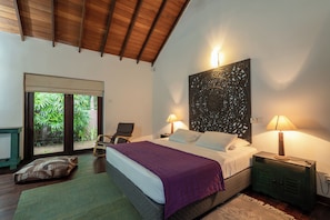 A bedroom with a bed, two bedside lamps, a nightstand, a chair, a window with a view of greenery, and a decorative wall piece. - The Aluthgama House by TSC (Dharga Town)