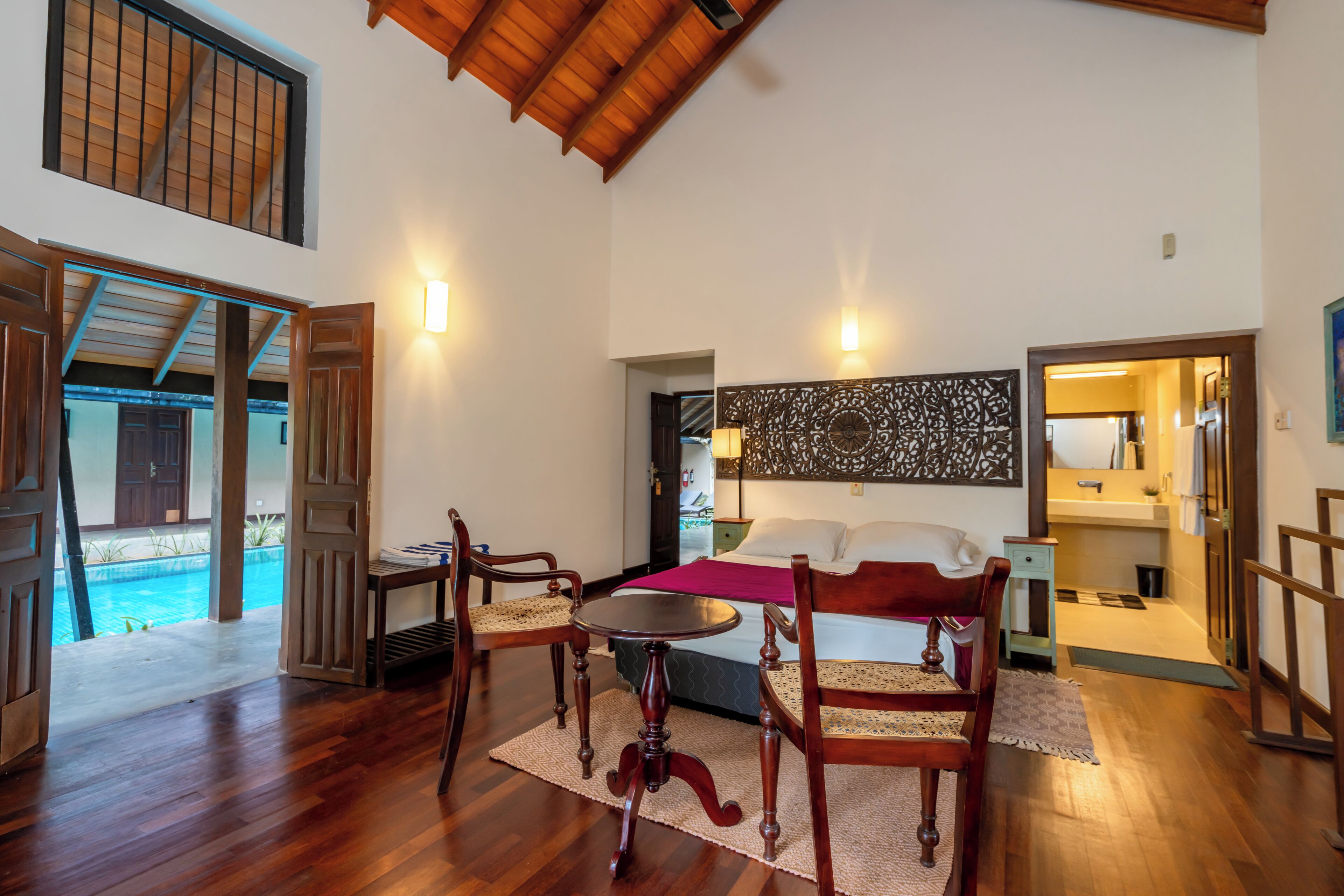 Una habitación amplia con cama, una mesita, dos sillas, un panel decorativo y vistas a una piscina.