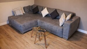 Un sofá seccional gris con cojines estampados, una mesa de centro de vidrio y un control remoto, y piso de madera.