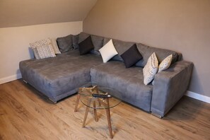Ein graues Ecksofa mit gemusterten Kissen, ein Glastisch mit Fernbedienung und Holzboden.