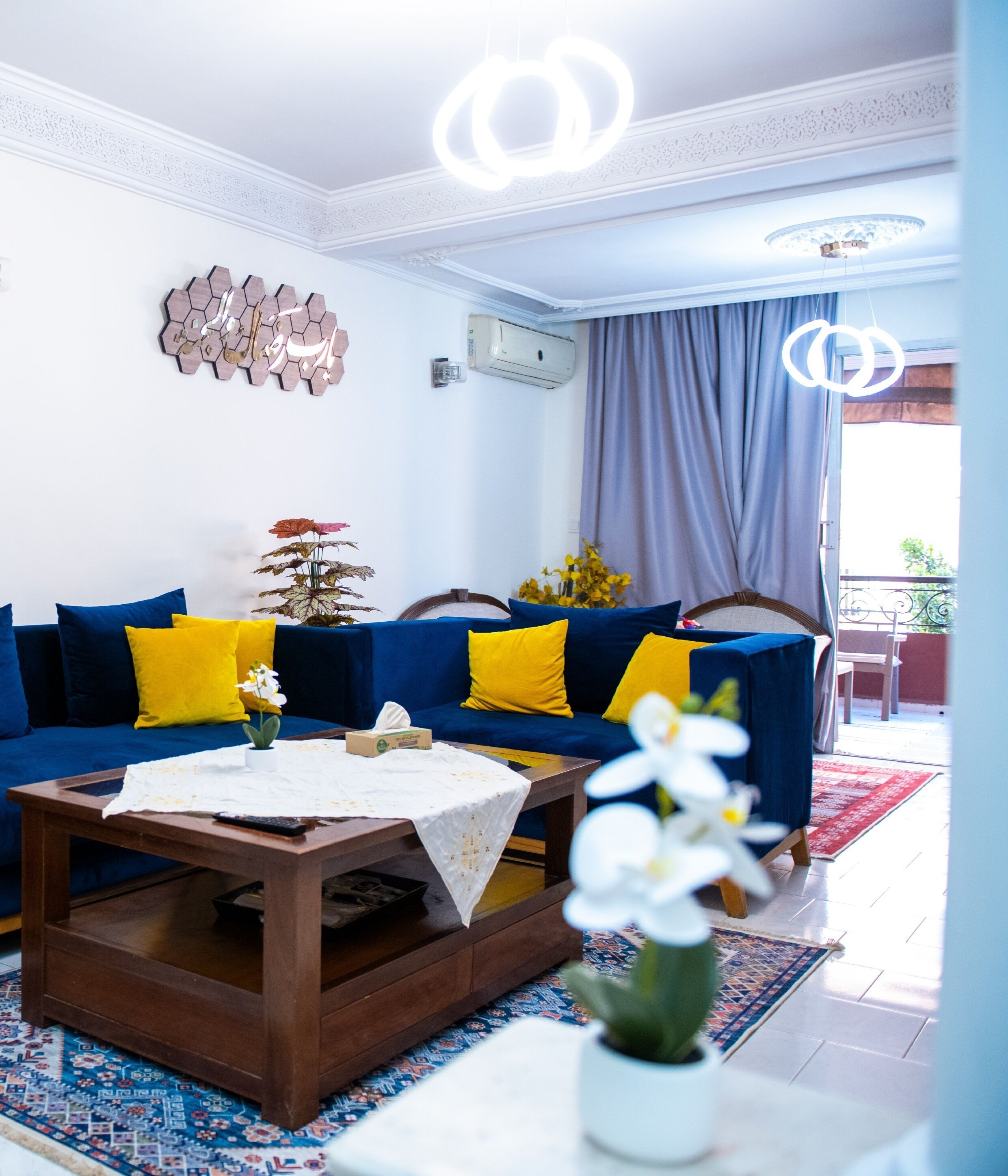 Un salon avec un canapé bleu, des coussins jaunes, une table basse en bois et un vase blanc rempli de fleurs.