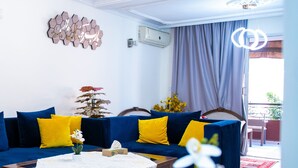 Una sala de estar con un sofá azul, cojines amarillos, una mesa de centro de madera y un florero blanco con flores.