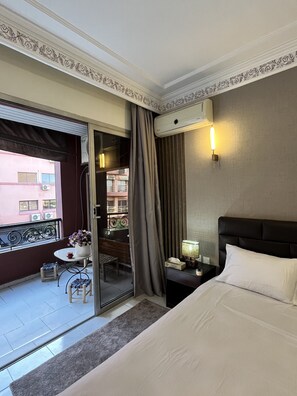 Ein Hotelzimmer mit Bett, Nachttisch, Lampe, einem kleinen Tisch mit Blumenvase, Stuhl und Blick auf den Balkon.