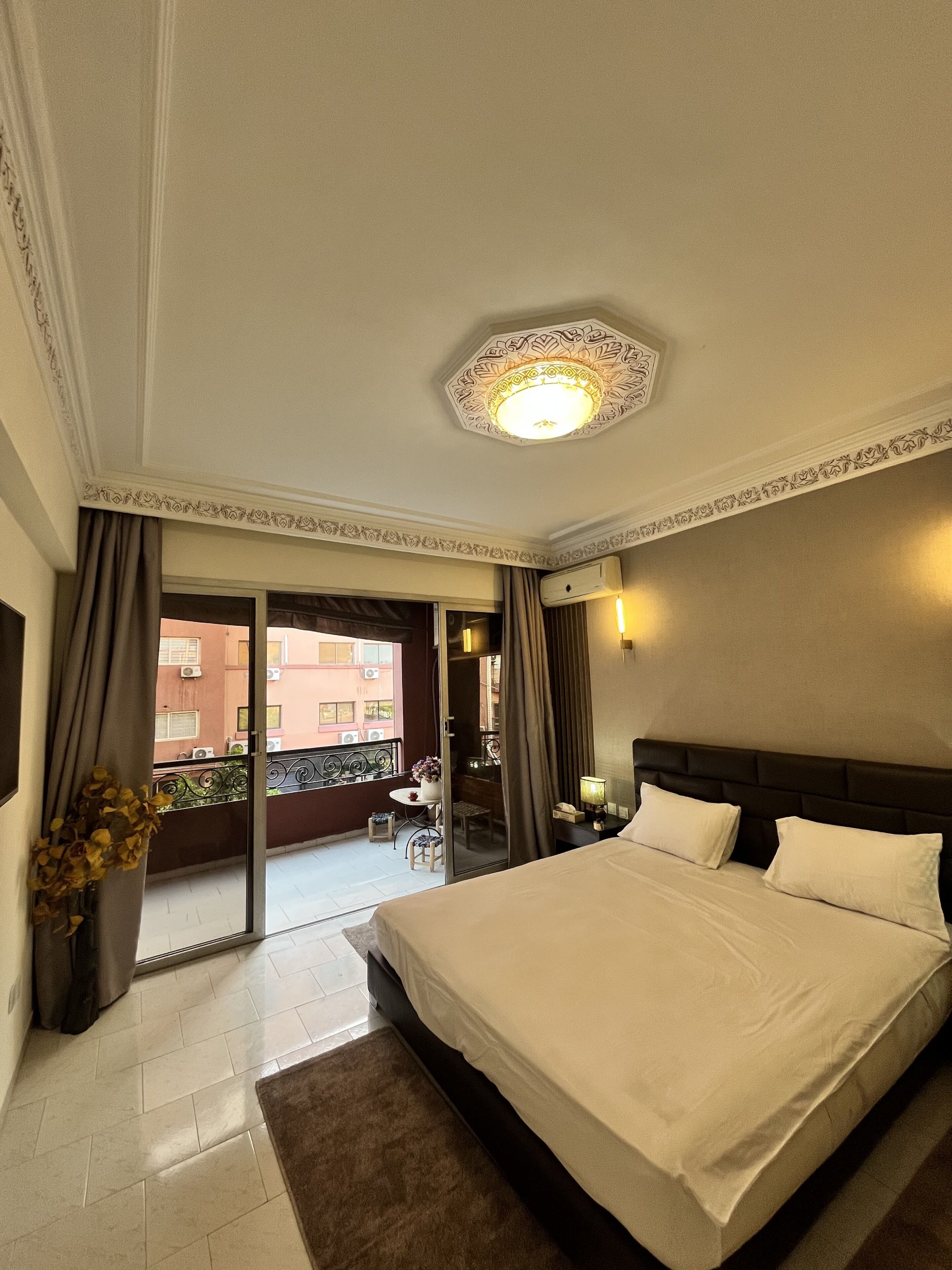 Une chambre d’hôtel avec un grand lit, un balcon donnant sur les immeubles et un plafond agrémenté d’un luminaire décoratif.