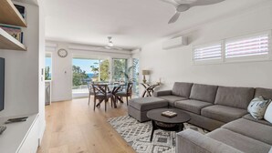 Una sala de estar moderna con un sofá modular, una mesa de comedor con sillas, un ventilador de techo y un ventanal con persianas.