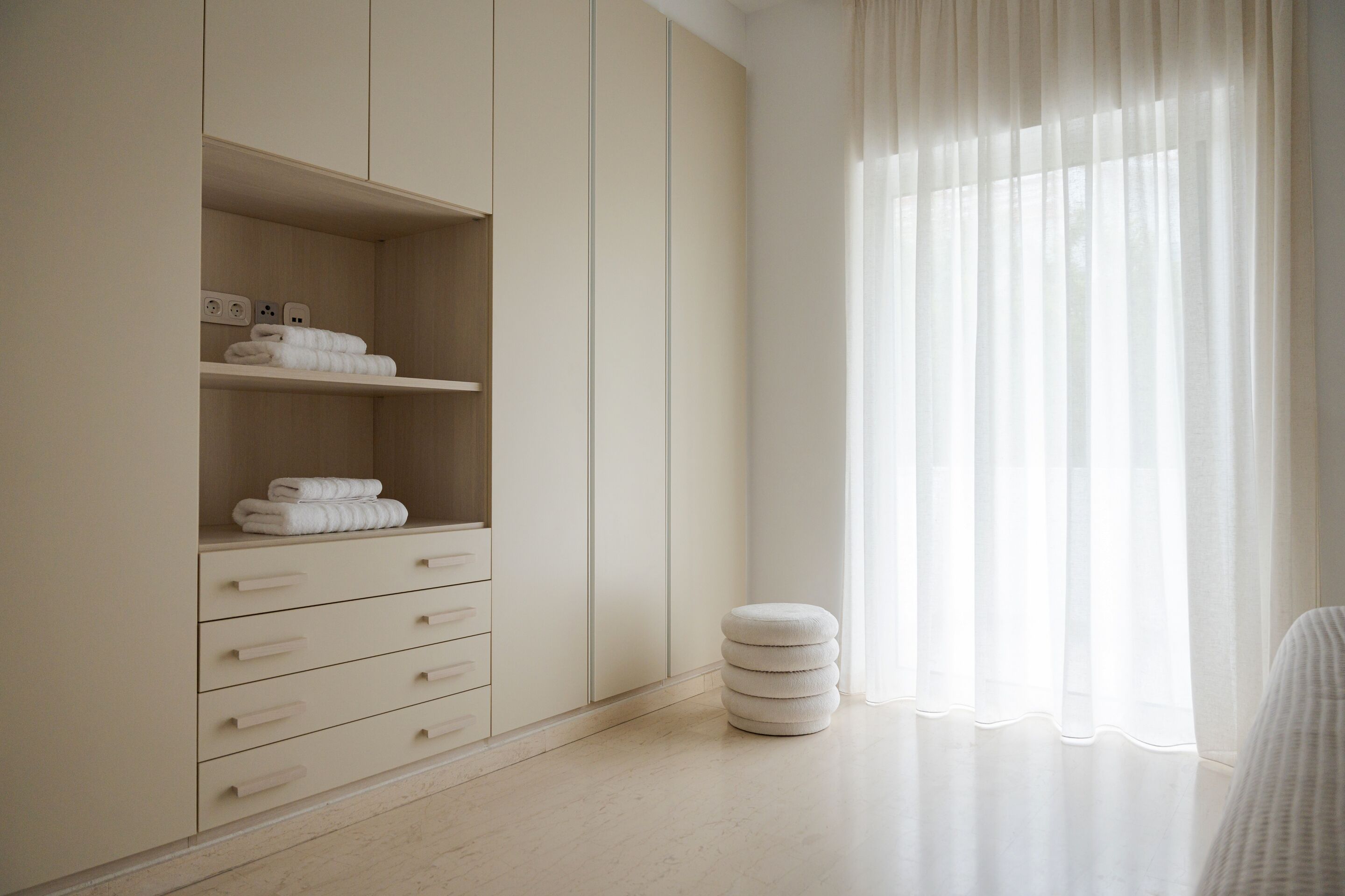A minimalist bathroom with a built-in wardrobe, a small stool, and a window with sheer curtains.