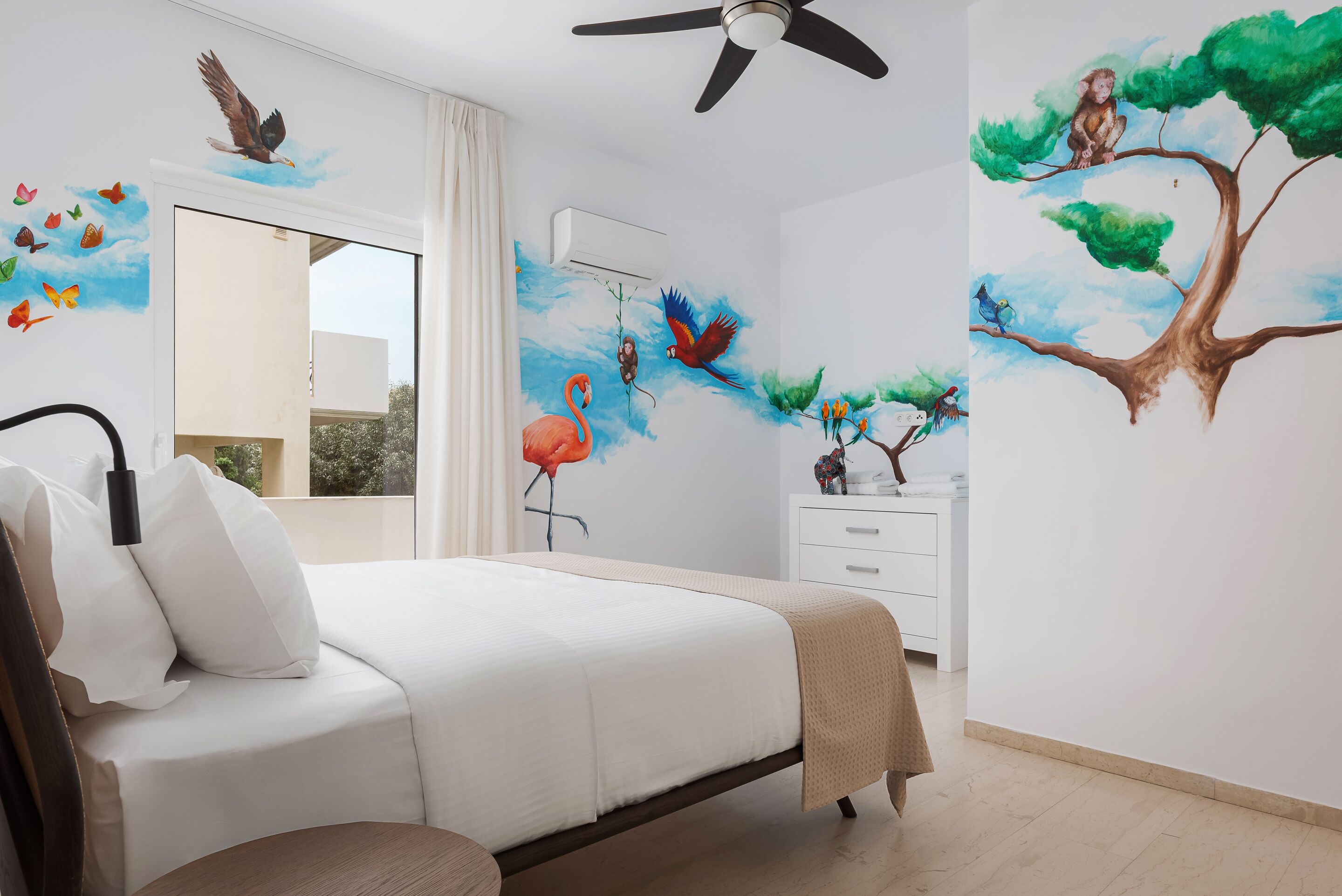 Un dormitorio con cama, ventilador de techo, mesita de noche, cómoda y un mural en la pared con pájaros y un árbol.