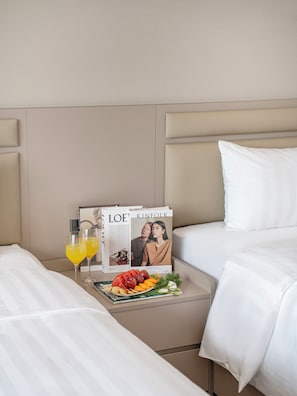 Quarto de hotel com cama, mesa de cabeceira com uma revista, dois copos de suco de laranja e um prato de frutas.