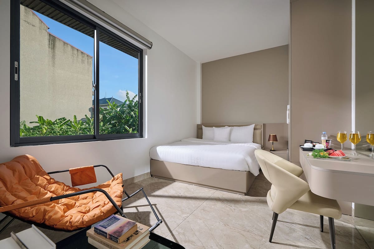 Quarto de hotel moderno com cama, escrivaninha com um laptop, cadeira e janela com vista para área verde.