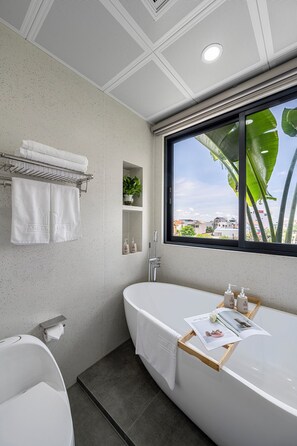 Um banheiro moderno com uma banheira livre, uma janela com vista para edifícios e vegetação, e um porta-toalhas com toalhas dobradas.