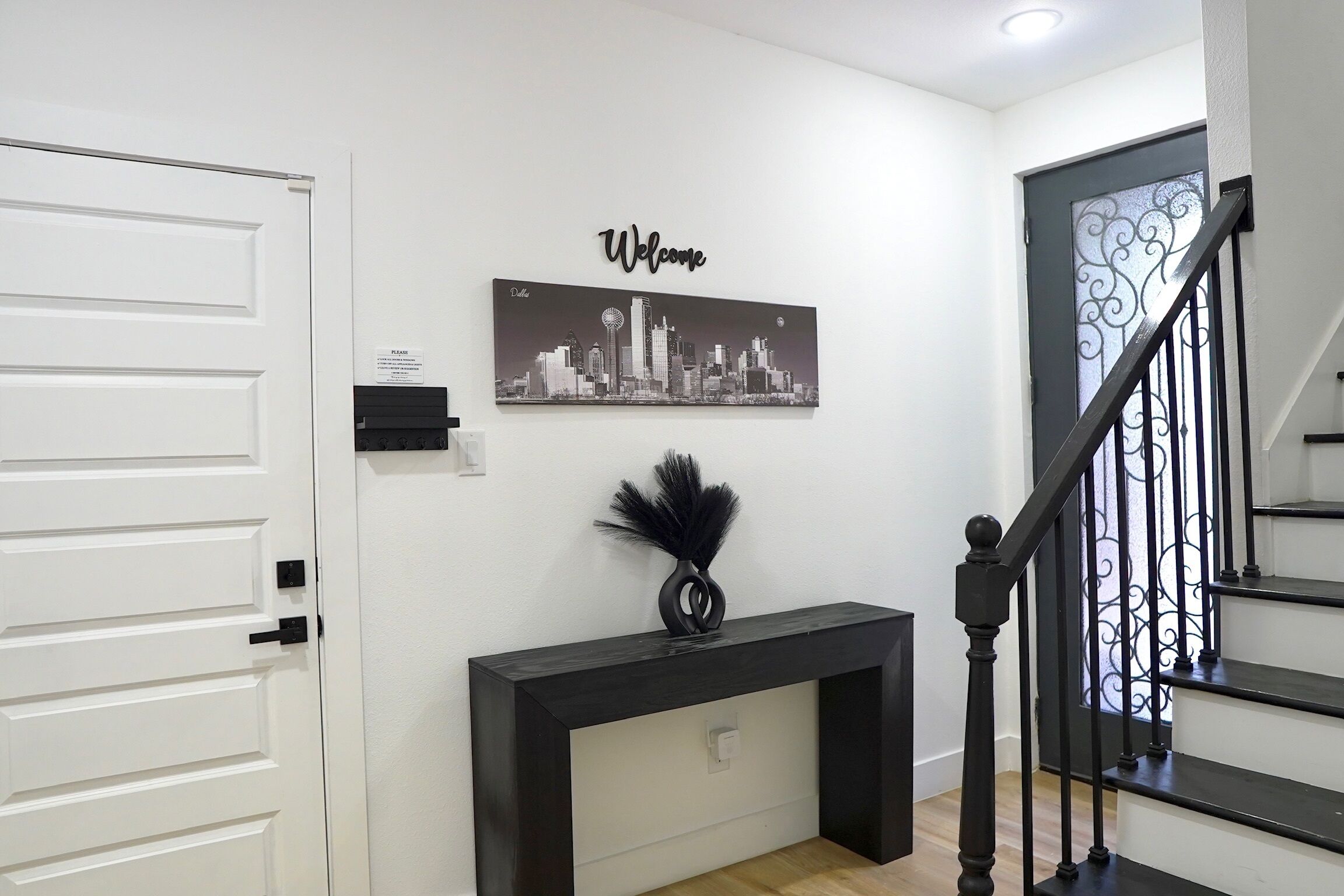 A hallway with a white door, a black console table with a black vase, a 'Welcome' sign, and a staircase with black railings.