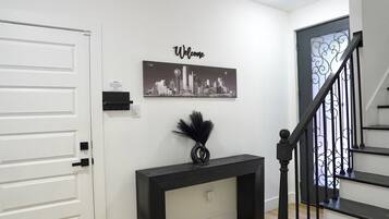 A hallway with a white door, a black console table with a black vase, a 'Welcome' sign, and a staircase with black railings.