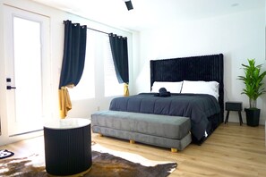 Ein Schlafzimmer mit einem Bett, einem kleinen Nachttisch, einer Topfpflanze, einem runden Hocker und einem Fenster mit Vorhängen.
