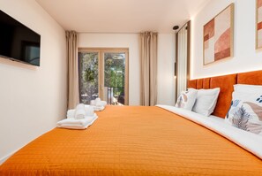 Ein Hotelzimmer mit einem großen Bett, einem Flachbildfernseher, einem Balkon mit Vorhängen und Blick auf Grünflächen.