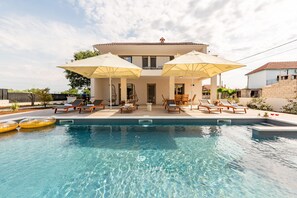 Ein Schwimmbecken mit Liegestühlen und Sonnenschirmen, ein Gebäude mit Balkon und ein klarer Himmel.