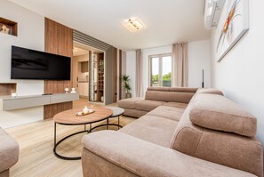Ein modernes Wohnzimmer mit Flachbildfernseher, einer Couch, einem Couchtisch und einer Schiebetür, die auf einen Balkon führt.