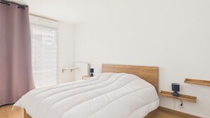 Un dormitorio con una cama grande, una mesita de noche con lámpara, un cabecero de madera, una ventana con cortinas y un estante en la pared.