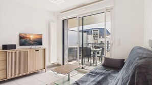 Una sala de estar moderna con una mesa de centro de vidrio, un sofá, un televisor sobre un mueble de madera, y un balcón con vistas a edificios.