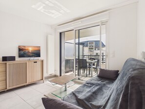 A modern living room with a glass coffee table, a sofa, a TV on a wooden cabinet, and a balcony with a view of buildings. - 950m from the beach, apartment for 2 (Le Pouliguen)