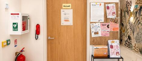 A reception area with a wooden door, emergency equipment, a fire extinguisher, and a bulletin board with various notices.