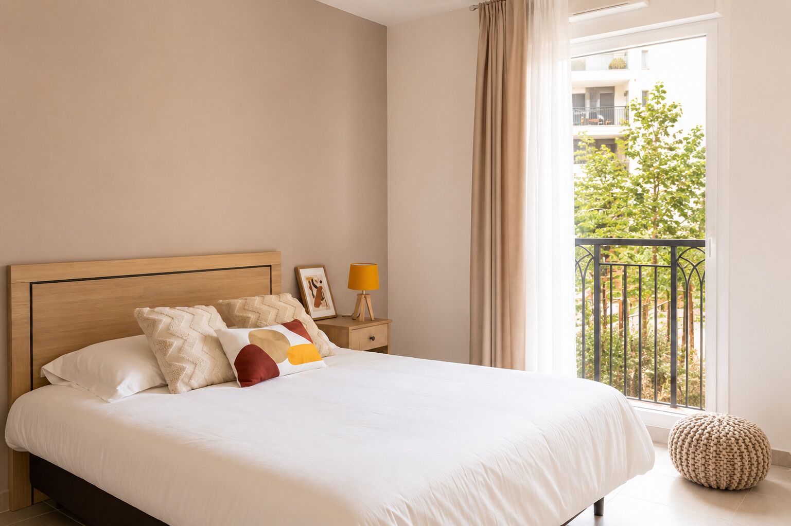 Une chambre à coucher comprenant un lit, une table de chevet, une lampe, une fenêtre donnant sur un balcon et un pouf rond.