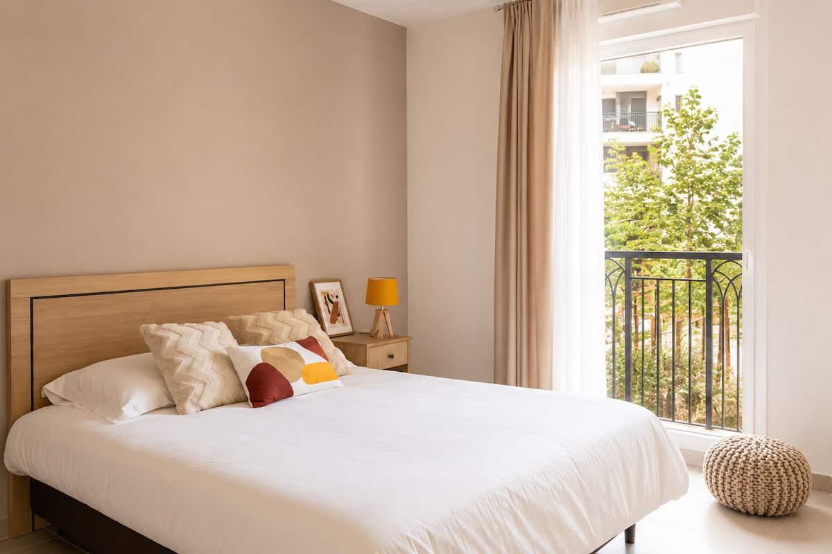 Une chambre à coucher comprenant un lit, une table de chevet, une lampe, une fenêtre donnant sur un balcon et un pouf rond.