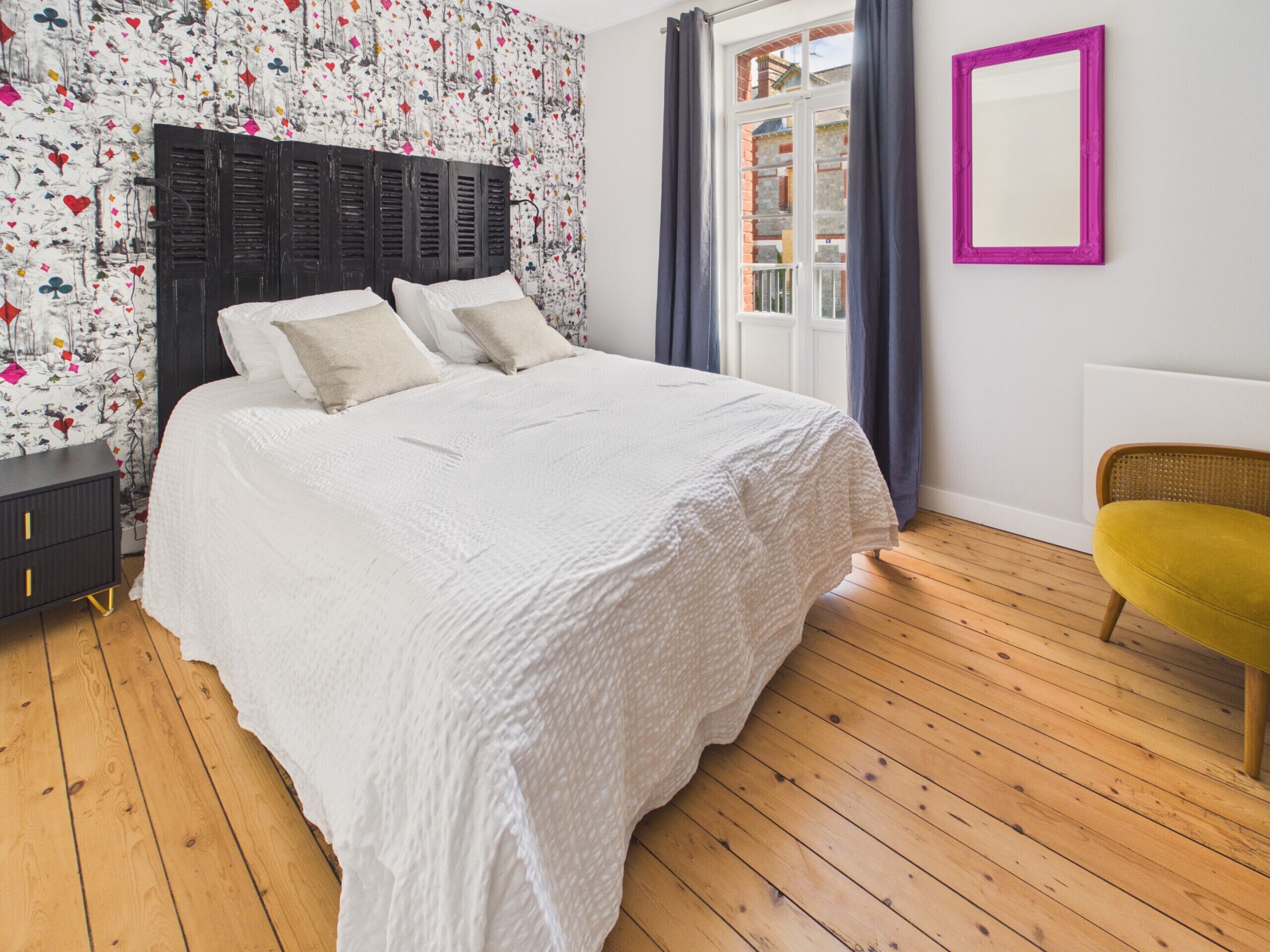 Ein Schlafzimmer mit Bett, Nachttisch, Stuhl, einem Fenster mit Vorhängen und einem Spiegel an der Wand.