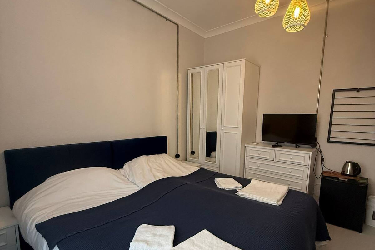 Una cama bien tendida con ropa de cama blanca y azul marino, una mesita de noche con un hervidor, un televisor sobre un mueble bajo y un armario con espejo.