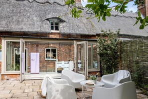 A thatched-roof building with a brick facade, a glass door entrance, and a patio with white furniture. - Plum Tree Cottage -  a cottage that sleeps 4 guests  in 2 bedrooms (Sudbourne)