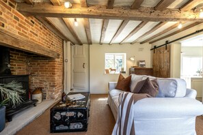 A rustic living room with exposed wooden beams, a brick fireplace, a white sofa, and a vintage suitcase. - Plum Tree Cottage -  a cottage that sleeps 4 guests  in 2 bedrooms (Sudbourne)