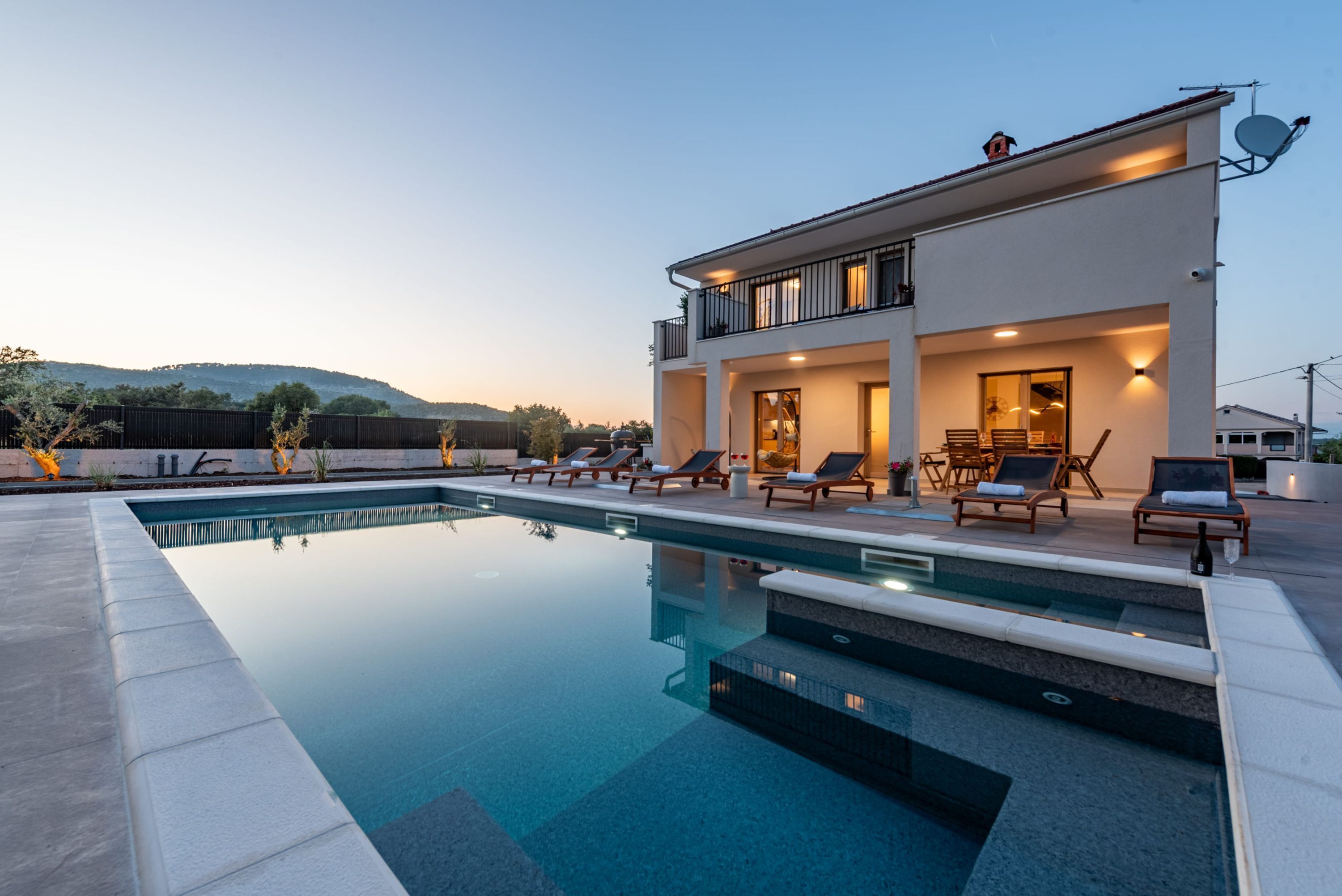 A modern two-story house with a swimming pool, outdoor seating, and a view of the hills.