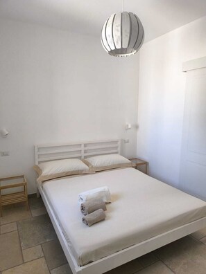 A single-bed room with a white headboard, two bedside tables, a hanging light fixture, and a door on the right. - The Ulivo Ritorto relaxes among ancient olive trees, just minutes from the beaches of Ostuni (Ostuni)