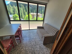 A room with a dining table, chairs, a sofa, and a large window offering a view of a forested area.