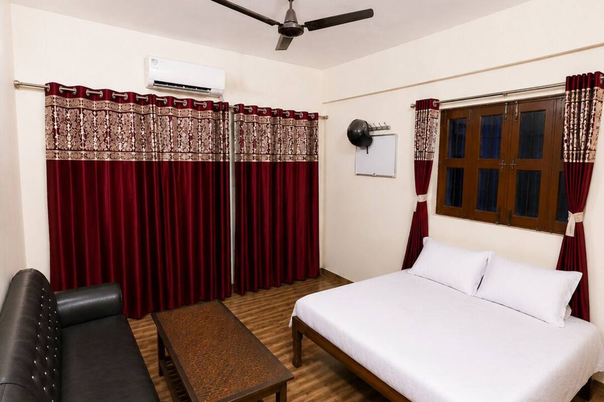 Habitación de hotel con cama, sofá de cuero negro, mesa de madera, ventilador de techo y ventana con cortinas.