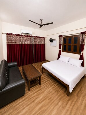 A hotel room with a bed, a black leather sofa, a wooden table, a ceiling fan, and a window with curtains. - Balaji Hotel (Farrukhabad)