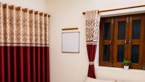 Un dormitorio con cama, cortinas, una ventana con contraventanas de madera, una pizarra blanca y una pequeña planta en maceta.