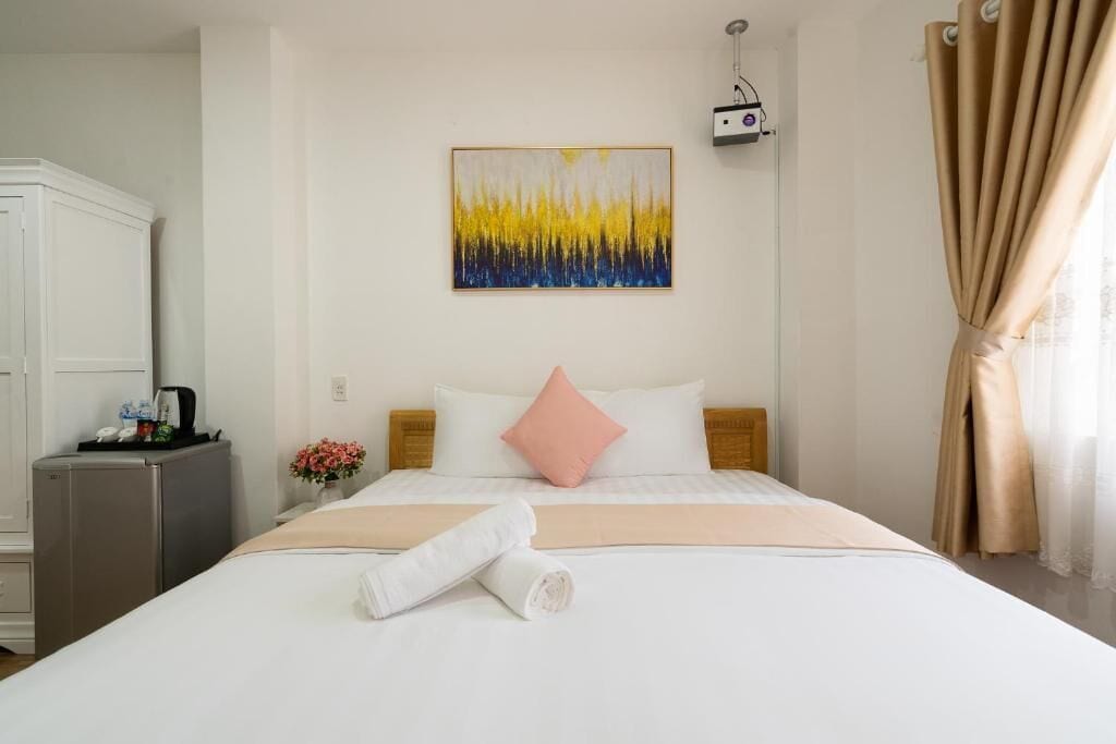 A hotel room with a bed, a small desk, a refrigerator, a painting on the wall, and a window with curtains.