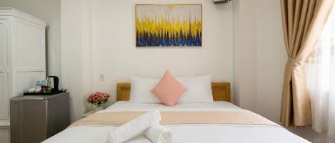 A hotel room with a bed, a small desk, a refrigerator, a painting on the wall, and a window with curtains.