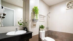 Casa de banho com lavatório, sanita e chuveiro. Há plantas na parede e um ventilador fixado na parede.