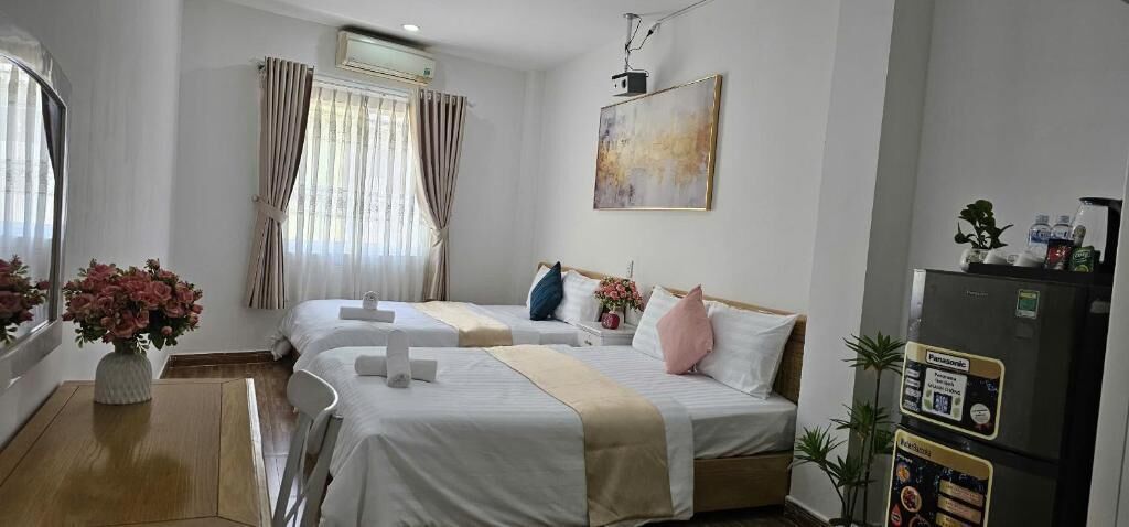 A hotel room with two beds, a wooden desk, a refrigerator, and a window with curtains.