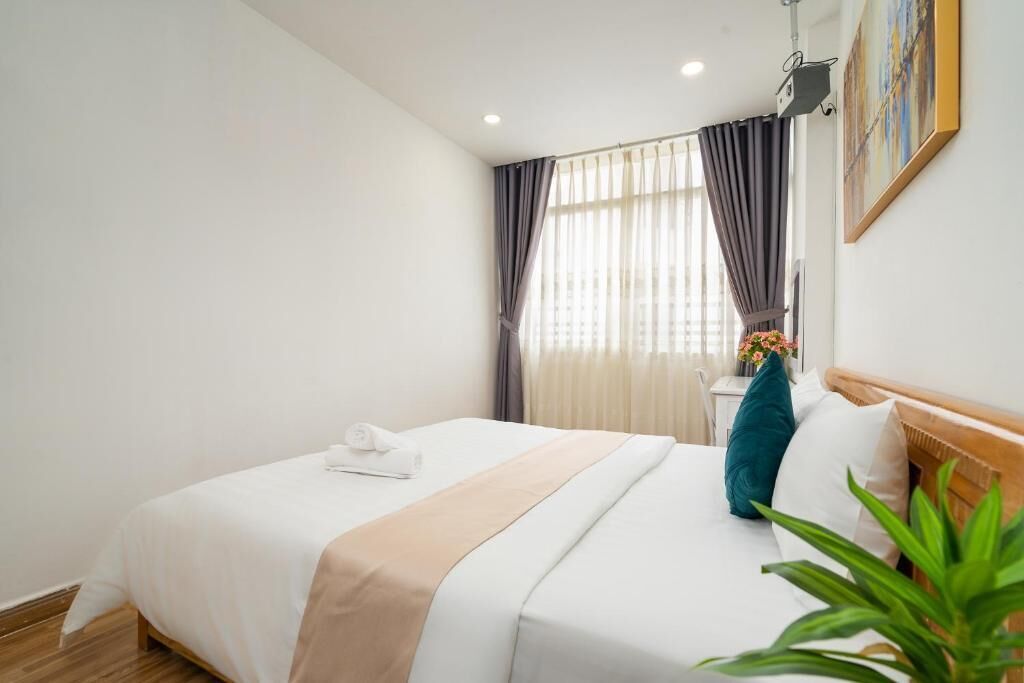 A hotel room with a bed, a bedside table, a window with curtains, and a plant.